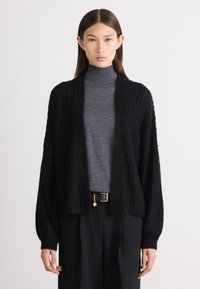 Črn pleten kardigan z odprtim sprednjim delom, rebrasto teksturo in dolgimi rokavi, oblečen čez sivo turtleneck majico in črne hlače, pritrjene z pasom.