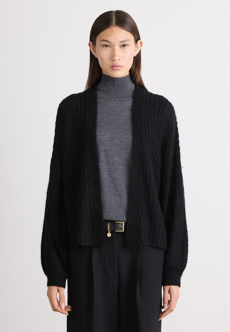 Črn pleten kardigan z odprtim sprednjim delom, rebrasto teksturo in dolgimi rokavi, oblečen čez sivo turtleneck majico in črne hlače, pritrjene z pasom.