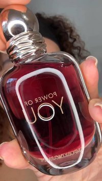 Dłoń trzymająca czerwony flakon perfum z napisem "Power of You", z srebrną kulistą nakrętką i metalową ramką.