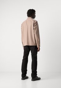 Lysbeige jakke med en glat tekstur, knapmanchetter og en subtil krave, parret med sorte bukser og sorte sko.