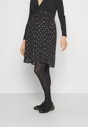 Frau trägt ein schwarzes Langarmoberteil, einen schwarzen Rock mit kleinen goldenen Punkten, schwarze Strumpfhosen und schwarze Ballerinas, steht vor einem schlichten Hintergrund.