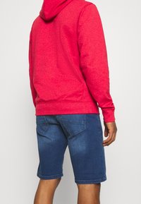 Sudadera con capucha de algodón rojo con cordón en la capucha, combinada con pantalones cortos de mezclilla azul oscuro que cuentan con dos bolsillos traseros y una textura descolorida.