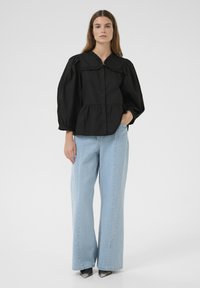 Zwarte top met pofmouwen en een gerimpelde kraag, knoopsluiting en tailleband, gecombineerd met lichtblauwe broek met brede pijpen en zwarte hakken.
