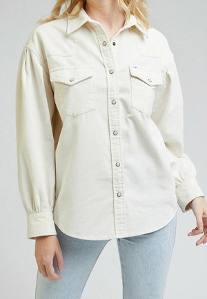 Femme portant une chemise en velours côtelé crème à boutons avec deux poches à rabat devant et un jean bleu clair, debout devant un fond uni.