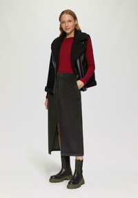 Gilet en simili cuir noir avec col en peau de mouton, haut bordeaux à manches longues, jupe en velours côtelé verte avec fente à l'avant, et bottes noires épaisses.
