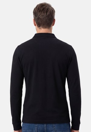 Mann mit kurzen braunen Haaren, der ein schlichtes schwarzes Langarmshirt und blaue Jeans trägt, steht mit dem Rücken zum Betrachter vor hellem Hintergrund.