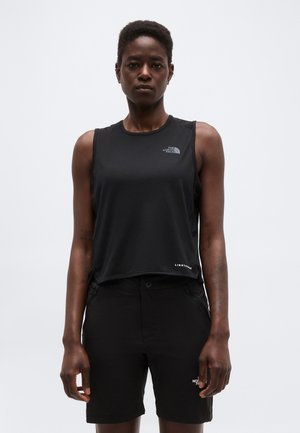 Frau trägt schwarzes ärmelloses Sporttop und passende Shorts, steht vor einem schlichten hellen Hintergrund.