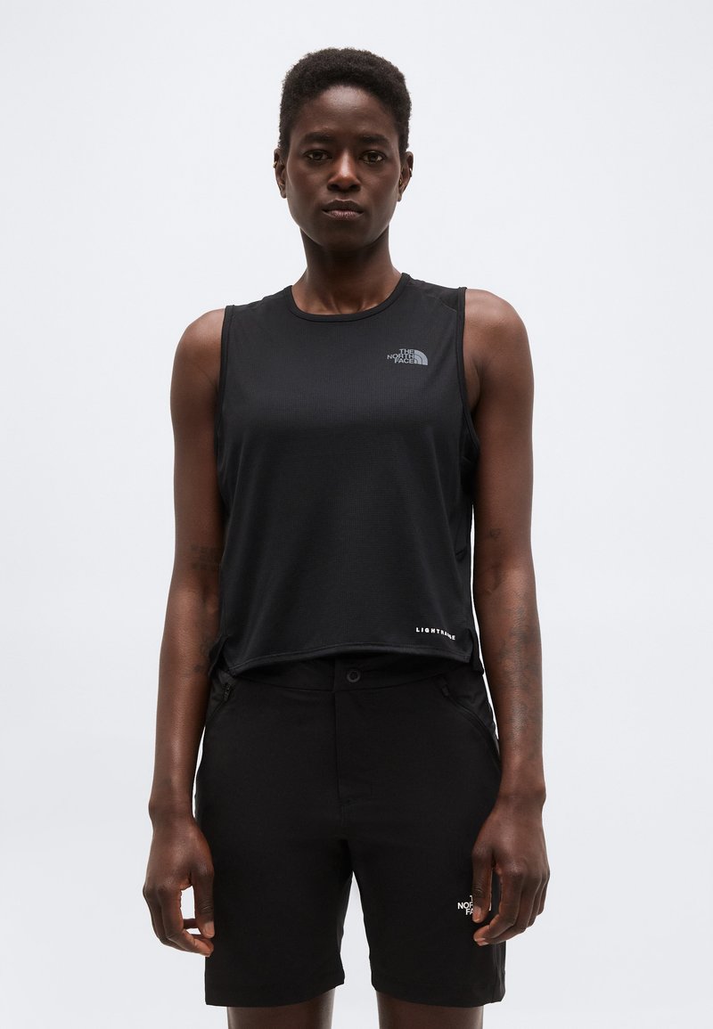 Γυναίκα με μαύρο αμάνικο αθλητικό τοπ και ταιριαστό σορτς, στέκεται μπροστά σε απλό ανοιχτό φόντο.