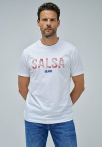 Hvid bomulds t-shirt med fed, rød og blå tekst "SALSA JEANS" med et strukturelt design, parret med blå denim jeans.