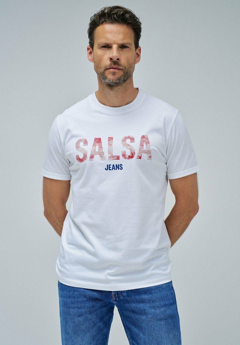 Camiseta blanca de algodón con un texto en rojo y azul en negrita "SALSA JEANS" con un diseño texturizado, combinada con unos jeans de mezclilla azul.