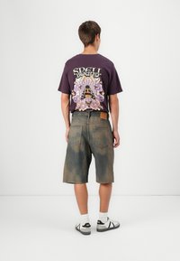 Purpura grafiskais T-krekls ar krāsainu dizainu aizmugurē, apvienots ar ceļa garuma nolietotām denim šortiem un melnbaltiem apaviem.