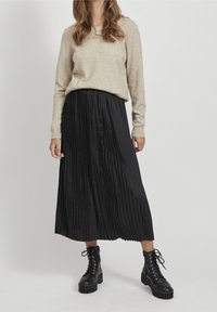 Beige tröja med långa ärmar i kombination med en svart plisserad midikjol och svarta snörande kängor. Slät tyg med texturerade plisseringar.