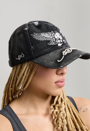 Woman with long blonde braids wearing a distressed black cap featuring a white winged skull embroidery and cursive script detail.