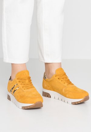 Personne portant des baskets jaune moutarde avec des semelles blanches et marron et des détails à motif serpent, associées à un pantalon blanc.