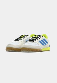 Bílé sportovní boty s modrými pruhy, černými tkaničkami a žlutým akcentem. Mají gumovou podrážku a svršek s semišovou texturou.