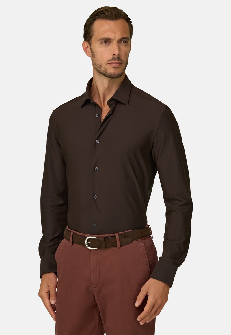 Homme portant une chemise ajustée marron foncé à manches longues boutonnée et un pantalon marron rougeâtre avec une ceinture en cuir marron foncé, debout.