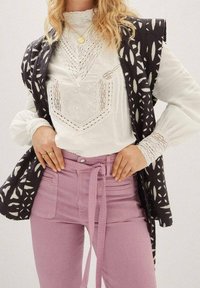 Czarny i biały wzorzysty kamizelka noszona na białej haftowanej bluzce, w połączeniu z różowymi spodniami z wysokim stanem i wiązanym paskiem.
