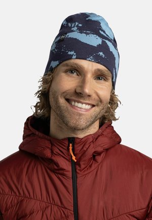 Knit beanie in navy with light blue abstract pattern. Soft texture, stretchy fit. Worn with a red puffer jacket featuring a zippered front.