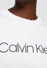Gros plan d'une personne portant un t-shirt blanc à col rond avec le texte noir « Calvin Klein » sur la poitrine.