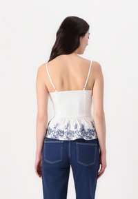Witte mouwloze top met smock-borstel, peplumrand met blauw bloemenpatroon. Gecombineerd met donkerblauwe jeans, met zicht op de achterkant.