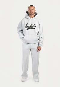 Sweatshirt gris clair à capuche avec logo "Smilodex" noir, associé à un pantalon gris assorti. Les deux pièces ont une coupe décontractée et une texture douce.