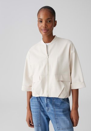 Femme portant une veste blanche à fermeture éclair avec poches à rabat et manches courtes retroussées, associée à un jean bleu taille haute.