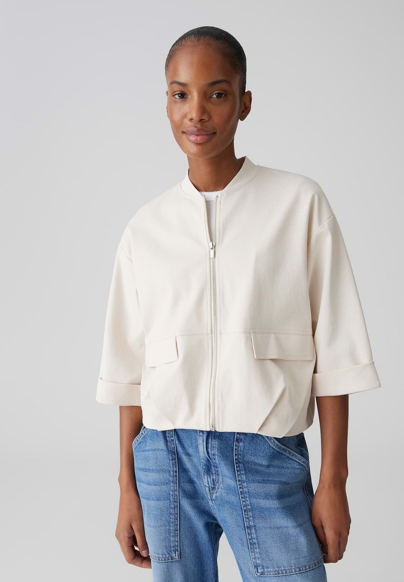 Vrouw draagt een wit jack met rits en klepzakken, korte mouwen met boord, gecombineerd met hooggesneden blauwe spijkerbroek.