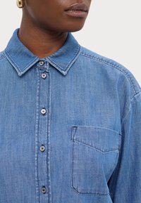 Ljusblå denimskjorta med spetskrage, framficka och synlig knapprad. Mjuk textur och avslappnad passform.