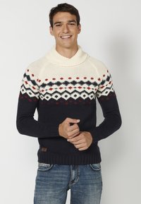 Suéter de punto azul marino con cuello de cisne crema, yugo estampado en rojo y blanco, y dobladillo acanalado. Llevado con jeans azules.