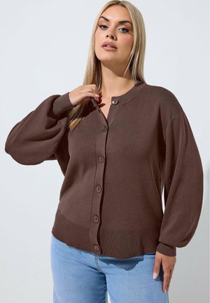 Vrouw met lang blond haar, gekleed in een bruine gebuttonneerde cardigan en lichtblauwe spijkerbroek, staand tegen een effen lichte achtergrond.