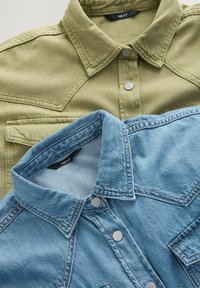 Twee over elkaar liggende overhemden: één in olijfgroen, de ander in lichtblauw denim. Beide hebben metalen drukknoopjes en borstzakken.