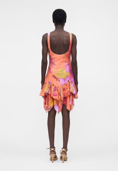 Γυναίκα με πορτοκαλί αμάνικο φόρεμα με floral μοτίβο και ασύμμετρο ρέλι με βολάν, φαίνεται από την πίσω πλευρά, σε συνδυασμό με μπεζ ψηλοτάκουνα σανδάλια.