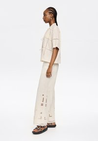 Beige top met korte mouwen en kantdetails, met een trekkoord bij de hals; bijpassende lichtcreme wijde broek met uitsnijdingen en bloembroderie.