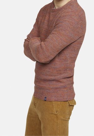 Persona con un suéter texturizado de color óxido, los brazos cruzados y pantalones de pana marrón claro frente a un fondo claro y liso.