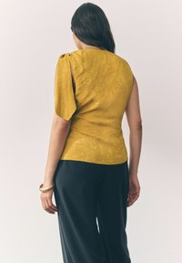 Geel top met een bladpatroon textuur, voorzien van een asymmetrische mouw en een slanke pasvorm, gecombineerd met zwarte broek en een gouden armband.