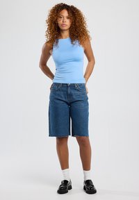 Vaaleansininen hihaton paita yhdistettynä polven pituisiin sinisiin farkkushortseihin, joissa on risat helmukset, yksinkertaista taustaa vasten. Mustat kengät ja valkoiset sukat.