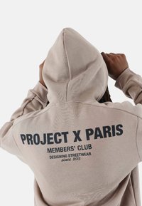 Sudadera con capucha beige con "PROJECT X PARIS" impreso en negro en la parte posterior, que presenta un ajuste relajado y un tejido texturizado para mayor comodidad.