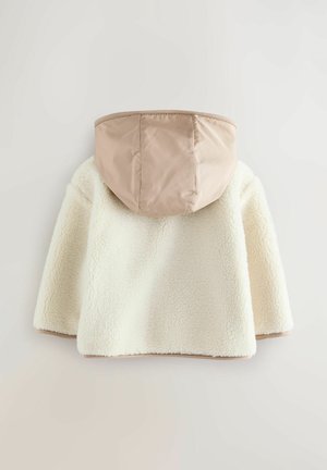 Krēmkrāsas flīsa jaka ar brūnganu kapuci, kas izceļas ar mīkstu tekstūru un noapaļotu formu, veidota siltumam un komfortam.