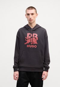 Mørkegrå oversized hettegenser i bomullsblanding, med et stort rødt grafisk logo med en okse og merkevarenavn. Frontlomme i kenguru-stil inkludert.