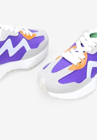 Zapatillas moradas con acentos grises, que cuentan con cordones blancos y detalles naranjas. Tejido suave con un diseño aerodinámico y suelas acolchadas.