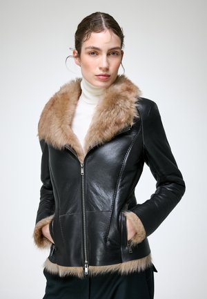Femme portant une veste en cuir noire avec un col et des poignets en fourrure beige, les mains dans les poches, et un pull à col roulé blanc en dessous.