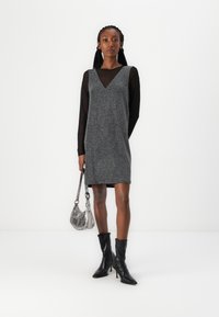 Vestido cinza sem mangas com decote em V sobre uma blusa preta de mangas longas, combinado com botas pretas até o tornozelo e uma bolsa transversal metálica.