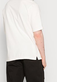 Vit bomullströja med korta ärmar, avslappnad passform och slits på sidan av fållen, bärs tillsammans med svarta byxor.