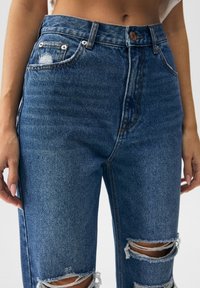 Vaqueros de mezclilla azul con cintura alta, rotos desgastados en las rodillas y herrajes de cobre. Presentan el clásico diseño de cinco bolsillos y una textura resistente.