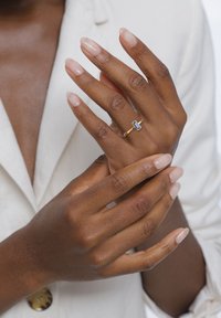 Hände mit dunklem Hautton, gut manikürten Nägeln, die einen goldenen Ring mit einem rechteckigen blauen Edelstein tragen, vor einem Hintergrund aus einem weißen Blazer.