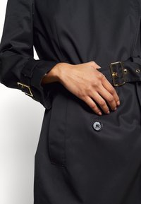 Czarny płaszcz trench z paskiem z złotą klamrą, zapięciem na guziki oraz ozdobnymi mankietami. Gładka tekstura i elegancki design z minimalnymi akcentami.