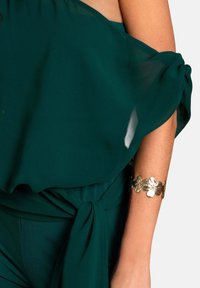Donkergroene, vloeiende top met een gestrikt taille-detail. Bevat doorschijnende stof en een gouden bloemenarmband om de arm. Zachte textuur en een elegante uitstraling.