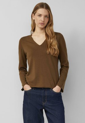 Jeune femme aux longs cheveux ondulés portant un haut marron à manches longues avec col en V et un jean bleu foncé, mains dans les poches, fond neutre.