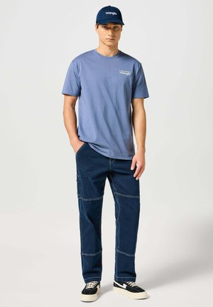 Blaues T-Shirt mit kurzen Ärmeln und Logo, kombiniert mit dunkelblauen Cargo-Hosen mit sichtbaren Nähten und schwarz-weißen Turnschuhen.