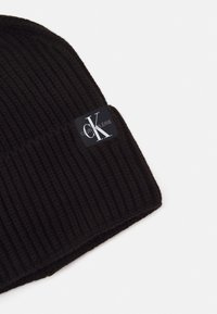 Gorro negro de punto con textura acanalada y un dobladillo plegado. Presenta un parche rectangular del logo de Calvin Klein Jeans en blanco y gris.